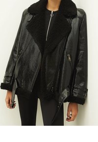 Czarna kurtka z eko skóry ze kołnierzem i mankietami z futerka, zamkami błyskawicznymi i luźnym krojem; ozdobiona dekoracyjnymi elementami metalowymi.