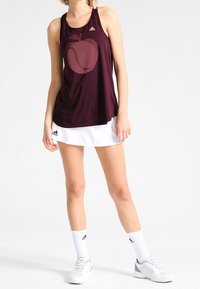 Débardeur de sport bordeaux avec un motif de pomme, associé à une jupe blanche. Présente un col rond et est fabriqué en tissu léger.