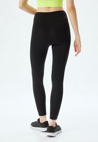 Zwarte leggings met een hoge tailleband, gemaakt van rekbaar materiaal. Kenmerkt zich door een gladde textuur en een strakke pasvorm, gecombineerd met zwarte sportschoenen.