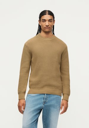 Giovane uomo con capelli lunghi intrecciati, indossa un maglione marrone lavorato a maglia e jeans azzurri chiari, in piedi su uno sfondo chiaro e semplice.
