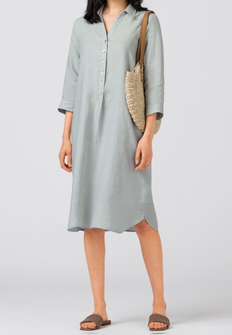 Kvinna som bär en ljusblå knäppbar midikjol, bär en vävd beige axelväska och har på sig bruna slidesandaler.