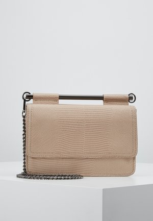 Bolso beige texturizado con asa de barra metálica y correa de cadena plateada, colocado sobre una superficie blanca frente a un fondo gris liso.