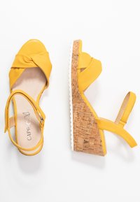 Gula kilklackar med en snodd strap-design, kork-texturerad klack och vit gummisula. Justerbar ankelrem för passform.