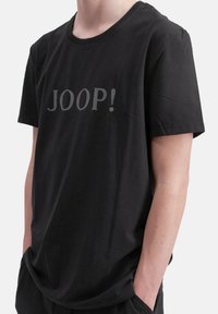 Camiseta de algodón negra con mangas cortas y cuello redondo, que presenta "JOOP!" impreso en gris en el pecho. Diseño simple y de corte relajado.
