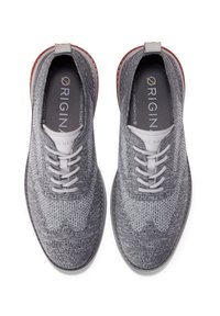 Zapatillas de punto grises con puntera redondeada, cordones blancos y acentos rojos contrastantes en el talón. Presentan una textura estampada en la parte superior.