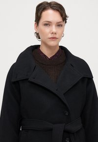 Melnas vilnas mētelis ar platu apkakli, priekšējiem pogām un jostu. Valkāts virs brūna teksturēta džempera ar sarkanu svītrotu krekla apkakli.