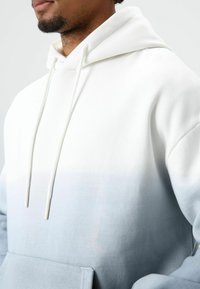 Sweatshirt met capuchon met een verloop van wit naar lichtblauw. Gemaakt van zachte stof met zichtbare stiknaden en een voorzak in kangaroostijl.