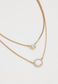 Collier à plusieurs couches en tone or avec deux chaînes. Une chaîne présente un pendentif en cristal rond, tandis que l'autre a un accent de pierre circulaire, orné de cristaux.