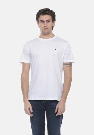 Uomo con capelli scuri che indossa una semplice maglietta bianca con un piccolo logo sul petto e jeans blu scuro, in piedi davanti a uno sfondo bianco.