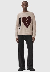 Άνδρας που φοράει μπεζ πλεκτό πουλόβερ με μαύρο γράμμα "I" και ένα σπασμένη κόκκινη καρδιά, συνδυασμένο με άνετο μαύρο τζιν και μαύρα παπούτσια.