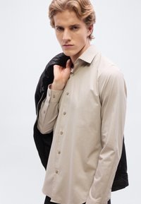Chemise beige clair à boutons avec manches longues, dotée d'un col classique et d'un léger éclat, portée sous une veste en cuir noire.