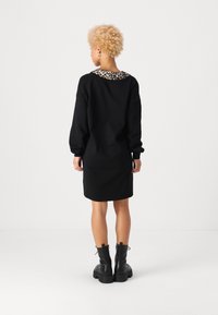 Črna obleka s kapuco in detajli z leopardjim vzorcem na ovratniku. Ima dolge rokave in sproščen kroj, v kombinaciji s črnimi debelimi čevlji.