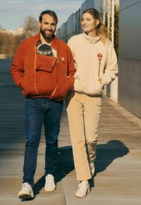Zwei Personen gehen auf einem Weg. Der Mann trägt eine orange Fleecejacke mit Kapuze; die Frau eine cremefarbene Fleecejacke. Beide in lässigen Hosen und Turnschuhen.