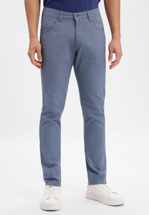 Mężczyzna w dopasowanych, teksturowanych niebieskich spodniach, białych tenisówkach i ciemnoniebieskiej koszuli, stojący na jednolitym tle.