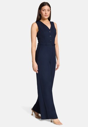 Femme debout portant une combinaison sans manches bleu marine avec trois boutons sur le devant et une ceinture assortie, associée à des sandales à talons transparents.