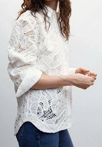 White lace blouse with intricate cut-out patterns, featuring a button-up collar and rolled sleeves. Worn with blue jeans.