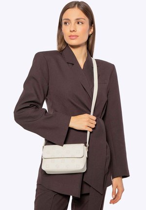 Žena v hnědém na míru šitém obleku držící na rameni bílou kabelku přes rameno s jemným květinovým vzorem.