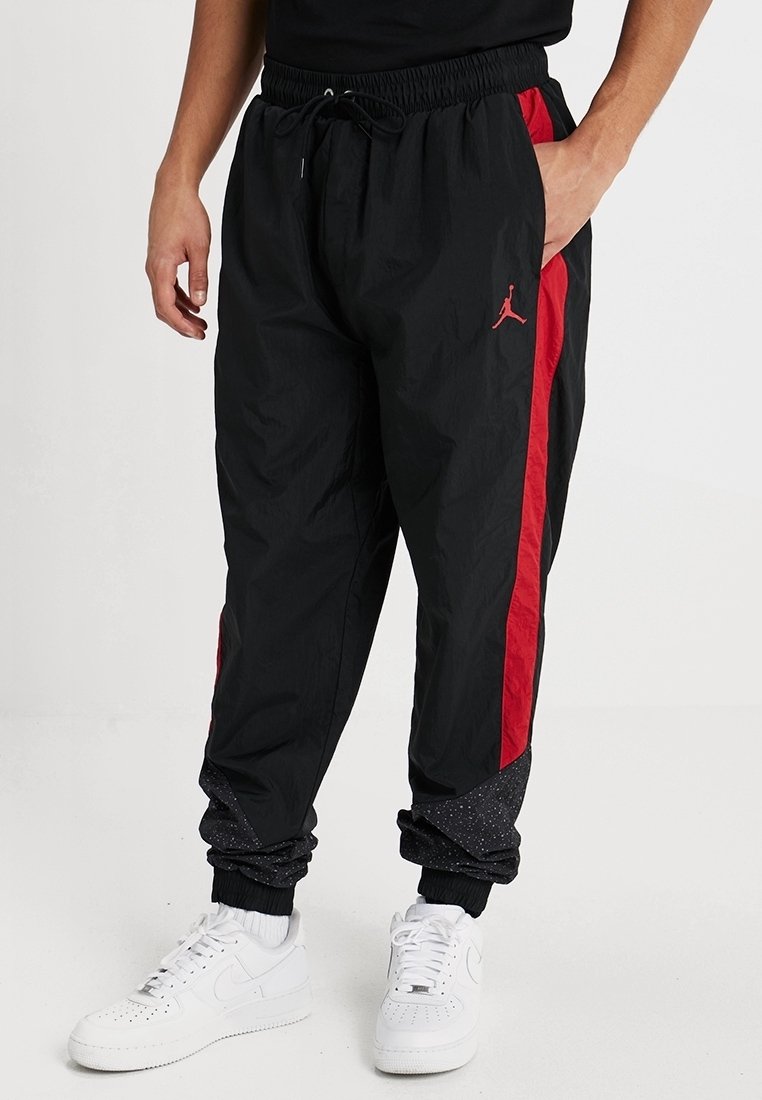 Svarta träningsbyxor med röda sidostråkar, elastisk midja och avsmalnande byxben med en texturerad svart detalj. Jordan-logotyp i röd.