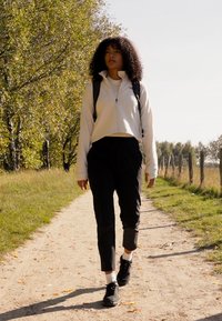 Veste blanche à zip, pantalon noir et baskets noires, avec un sac à dos. Cheveux bouclés, marchant sur un sentier en terre entouré d'arbres.