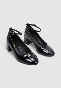 Chaussures à talons blocs en cuir verni noir avec une bride de cheville, bout rond et finition lisse. Logo doré à l'intérieur.