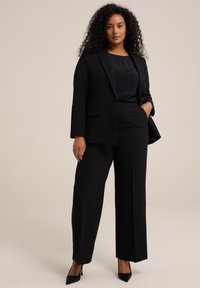 Costume noir ajusté comprenant une veste cintrée avec revers en pointes et un pantalon droit. Tissu lisse ; associé à des escarpins noirs à bout pointu.