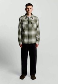 Camisa xadrez verde com dois bolsos no peito, design com colarinho e fecho de botão. Usada com calças pretas e sapatos castanhos escuros.