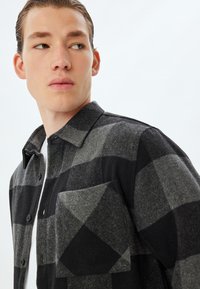Czarna i szara flanelowa koszula w kratkę z zapinaniem na guziki, kieszonką na piersi i kołnierzykiem. Materiał ma miękką, teksturowaną powierzchnię.