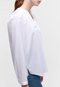 Chemise blanche à manches longues avec col en V, fentes latérales, texture lisse et coupe décontractée, associée à un jean en denim bleu.