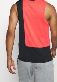 Débardeur sportif sans manches présentant un design en blocs de couleur avec un devant corail vif et un dos noir uni, fabriqué en tissu respirant.