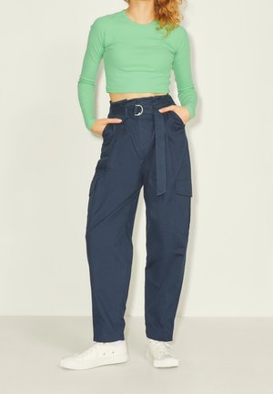 Eine Frau trägt ein grünes langärmliges Crop-Top, navyfarbene, hoch taillierte Hose mit Gürtel und Taschen sowie weiße Sneakers, während sie mit den Händen in den Taschen steht.