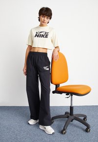 T-shirt Nike crema tagliata con logo nero; pantaloni cargo neri; sneakers Nike bianche. Sedia ergonomica arancione con schienale arrotondato.