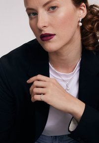 Srebrna biżuteria obejmuje naszyjnik z perłą w kształcie kropli, smukły pierścionek z małymi kamieniami oraz eleganckie kolczyki. Modelka ma na sobie czarną marynarkę i białą bluzkę.