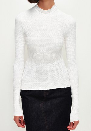 Hvid langærmet top med en tekstureret overflade og høj hals, der har en bølget kant og tætsiddende design, parret med en mørk denimnederdel.