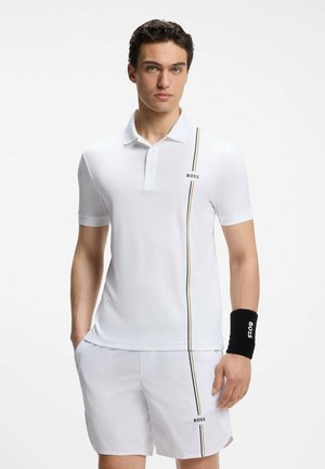 Jeune homme portant un polo blanc et un short avec une bande verticale et le logo "BOSS", bracelet noir au poignet droit.