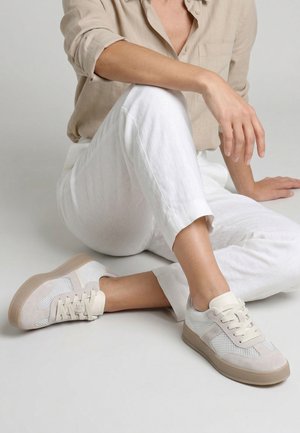 Osoba w beżowej koszuli i białych spodniach siedząca na podłodze, nosząca beżowo-białe siateczkowe trampki z gumową podeszwą.