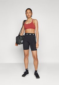 Rode sportbeha, zwarte high-waisted shorts met drie witte strepen op de tailleband, zwarte sneakers, terwijl een zwarte sporttas wordt vastgehouden.