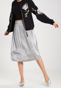 Svart bomberjacka med blommig broderi, ihop med en glänsande silverfärgad plisserad midi-kjol och silverfärgade spetsiga flats.