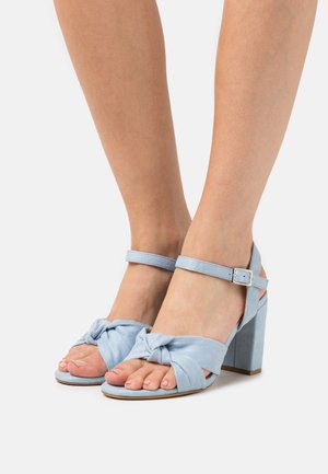 Světle modré sandály na podpatku s uzlovým designem na přední straně, kotníkovým páskem s kovovou sponou a čtveratým blokovým podpatkem. Měkká surová textura.