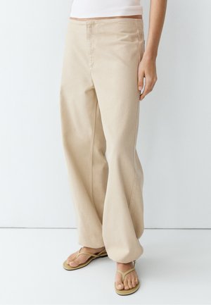 Persoon draagt losse beige broek en beige teenslippers, staand tegen een effen witte achtergrond.
