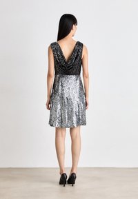 Abito nero e argento con paillettes, scollatura a cowl, senza maniche, silhouette svasata, apertura sul retro, tessuto testurizzato, abbinato a tacchi neri.