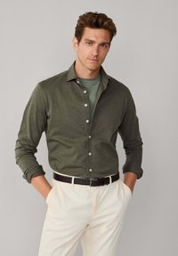 Uomo che indossa una camicia verde oliva abbottonata sopra una t-shirt verde, pantaloni beige e cintura nera, in piedi con le mani nelle tasche davanti a uno sfondo grigio.