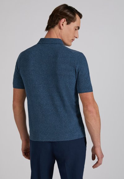 Mężczyzna stojący tyłem, ubrany w krótkim rękawem niebieską dzianinową koszulkę polo i ciemnoniebieskie spodnie na jednolitym tle.