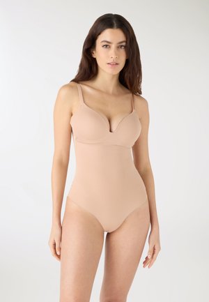 Donna con capelli lunghi castani che indossa una tutina aderente color nudo con spalline sottili su uno sfondo bianco uniforme.