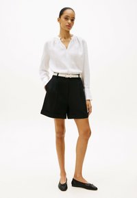 Blusa de lino blanca con escote en V y cuello fruncido, combinada con pantalones cortos negros a medida con dobladillo doblado y bolsillos frontales. Zapatos planos negros.