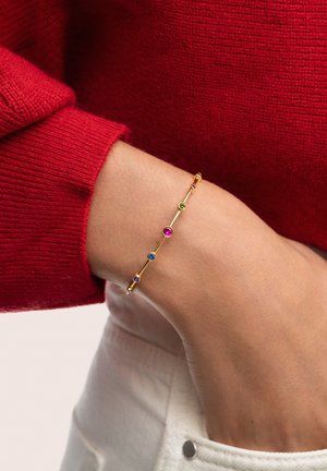 Pulsera de oro con cinco piedras multicolores: verde, rosa, morado y azul. El diseño es delgado y delicado, se lleva en la muñeca.