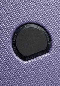 Materiale testurizzato viola con un distintivo nero circolare che mostra "Delsey Paris 1946." L'illuminazione mette in evidenza il motivo e i dettagli del logo.