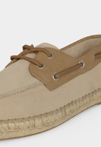 Μπεζ και ταμπελί παπούτσι τύπου boat κατασκευασμένο από σουέντ με δερμάτινα κορδόνια περασμένα μέσα από μεταλλικά κρίκια και πλεκτή σόλα από γιούτα.