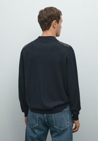 Muž ve tmavém svetr s dlouhými rukávy a šedými nášivkami na ramenou, v modrých džínách, stojí zády k divákovi proti prosté zdi.