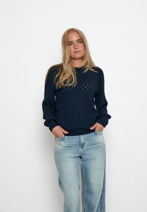 Donna bionda con maglione blu navy strutturato e jeans azzurri chiari in piedi con le mani nelle tasche davanti a uno sfondo bianco semplice.