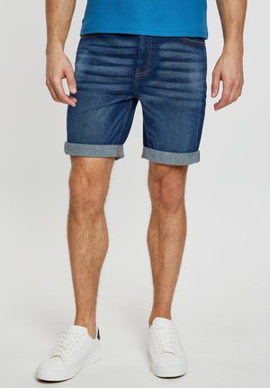 Pantalones cortos de mezclilla en azul oscuro con una textura suave, puños doblados y costuras marrones en contraste, combinados con zapatillas blancas.